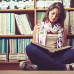 Full length of a female student sitting against bookshelf and re